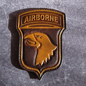 以下が含まれることがあります： 上部に「AIRBORNE」の文字が入った茶色とタン色の革製パッチ。パッチの中央には、詳細なワシの頭のデザインが施されています。縁は対照的な糸でステッチされています。