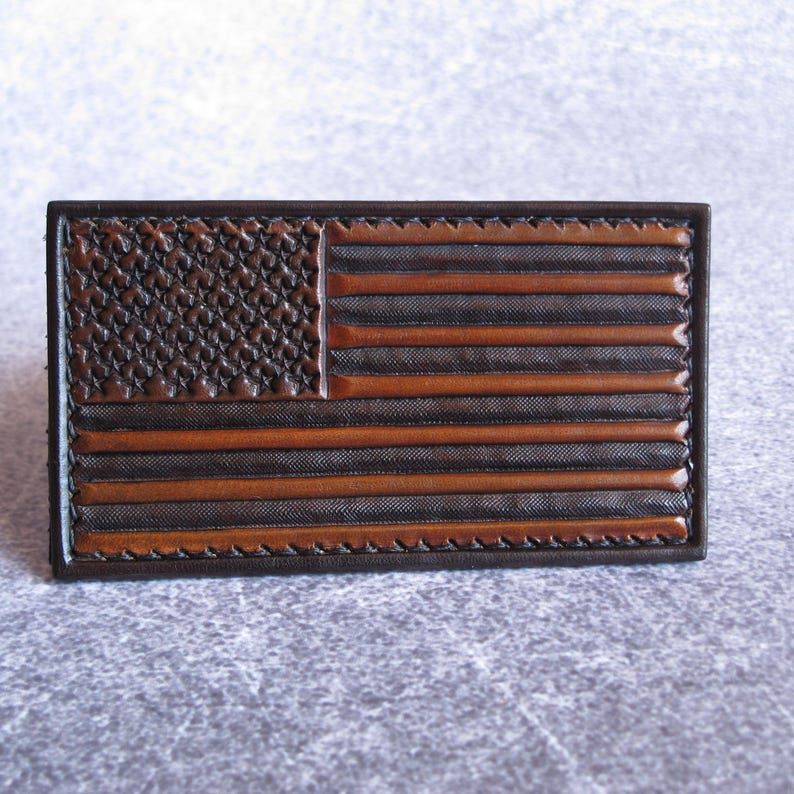 May include: A rectangular leather patch featuring an embossed American flag design. The flag displays alternating dark brown and tan horizontal stripes with a textured canton area containing embossed stars, all bordered by a stitched leather frame.