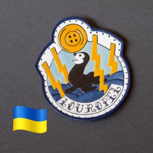 May include: A blue and yellow leather patch with a white background. The patch features a cartoon sea otter holding a yellow button with four holes. The text "OUROPE" is written in a white cursive font on the patch.
