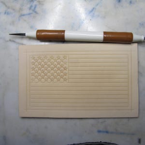 May include: A rectangular piece of light tan leather featuring a stamped American flag design with horizontal stripes and a field of stars. A white and brown stylus tool with a pointed metal tip is positioned horizontally above the leather piece.