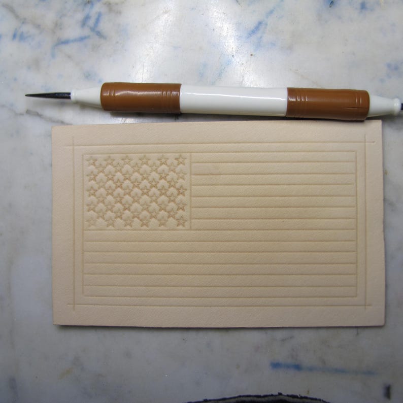May include: A rectangular piece of light tan leather featuring a stamped American flag design with horizontal stripes and a field of stars. A white and brown stylus tool with a pointed metal tip is positioned horizontally above the leather piece.