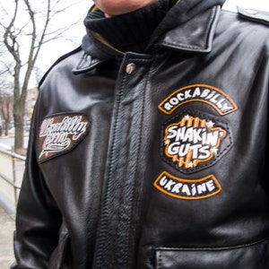May include: Black leather jacket with a zipper closure and multiple patches. The patches feature the text "Rockabilly Crew", "Shakin' Guts", and "Ukraine".