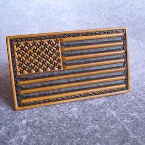 May include: A rectangular leather patch featuring a debossed American flag design. The piece displays alternating tan and dark brown horizontal stripes with a field of stars in the upper left corner, all framed by a decorative stitched border.