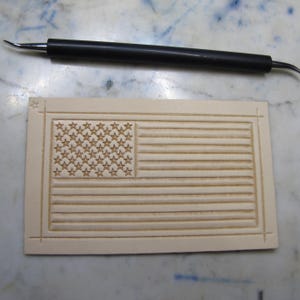 May include: A rectangular piece of light beige leather featuring a stamped American flag design with fifty stars and thirteen horizontal stripes, accompanied by a black metal stylus tool with a pointed tip for leather crafting and detailing.