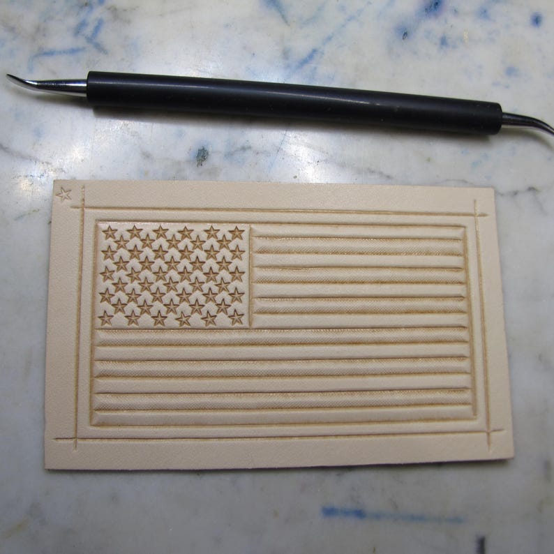 May include: A rectangular piece of light beige leather featuring a stamped American flag design with fifty stars and thirteen horizontal stripes, accompanied by a black metal stylus tool with a pointed tip for leather crafting and detailing.