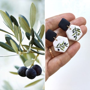 Può includere: L'immagine presenta un rametto di ulivo con olive e un paio di orecchini fatti a mano. Gli orecchini hanno la parte superiore quadrata nera e la parte inferiore esagonale bianca decorata con motivi di rami di ulivo.
