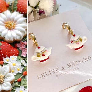 Puede incluir: Un par de pendientes de aro dorados con un dije de tetera blanca. La tetera tiene una tapa roja, hojas verdes y flores rosas. Los pendientes se muestran en una tarjeta blanca con el texto "CELINE & MASSIMO DEUTSCHLAND".