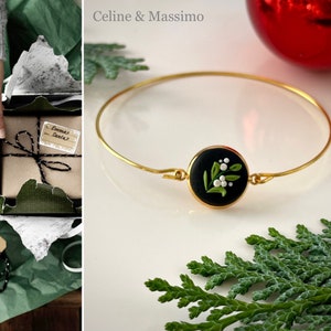 May include: A gold bangle bracelet with a black and white holly berry charm. The charm is made of polymer clay and features green leaves and white berries.