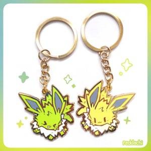 Puede incluir: Dos llaveros con personajes de dibujos animados. Cada llavero tiene un anillo y una cadena dorados. Los amuletos tienen la forma de un personaje de dibujos animados con acentos amarillos, verdes y blancos. El fondo es blanco con formas de estrellas verdes.