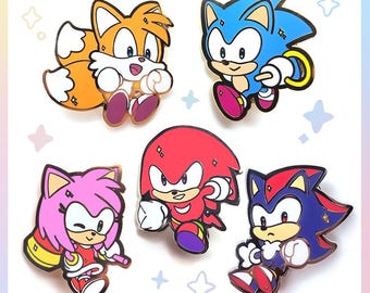 May include: Set of five enamel pins featuring characters from the Sonic the Hedgehog video game series. The pins include Sonic, Tails, Amy Rose, Knuckles, and Shadow, each in vibrant colors with gold-colored metal borders.