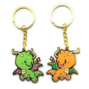 May include: Two keyrings with cartoon dragon charms. One is green with purple wings, the other is orange with green wings. Both have gold-coloured chains and key rings. The dragons have smiling faces and star accents.