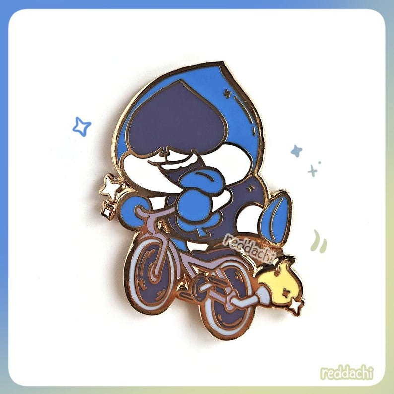 May include: A pin featuring a cartoon character riding a bicycle. The character is primarily blue and white with a gold outline. The bicycle is also gold and blue, with a yellow flame and star detail. The pin has the text "redddachi".