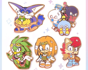 以下が含まれることがあります： ビデオゲームフランチャイズのキャラクターをフィーチャーした、カラフルなエナメルピンのコレクション。ピンには、カエルを抱えた紫と青のキャラクターと、ゴーグルをかけた緑色の鳥のキャラクターが含まれています。ピンには金色の縁取りが施されています。