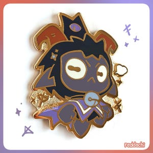 May include: A gold-colored enamel pin featuring a cartoon character with a dark blue face, purple accents, and a crown. The pin has a gold border and the text "reddachi" is visible.