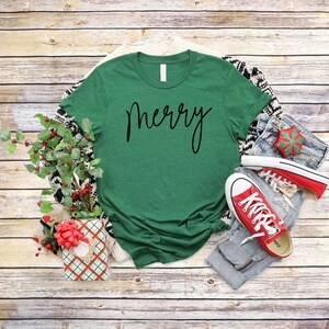 May include: A green t-shirt with the word "merry" written in black cursive script. The shirt is laid flat on a wooden surface with a pair of red sneakers, a pair of jeans, and a wrapped gift box.