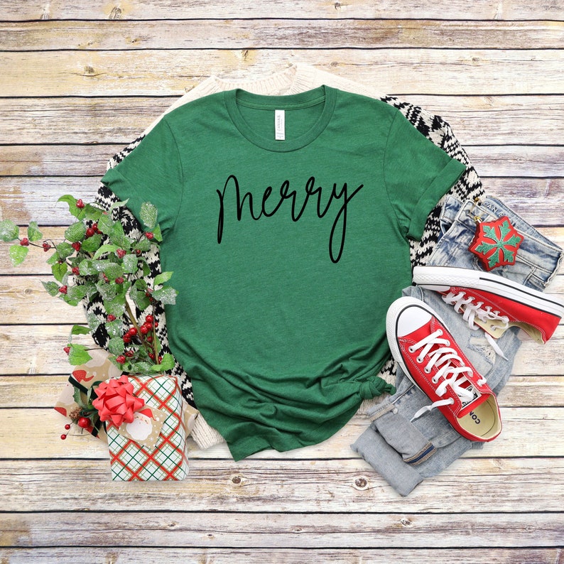 May include: A green t-shirt with the word "merry" written in black cursive script. The shirt is laid flat on a wooden surface with a pair of red sneakers, a pair of jeans, and a wrapped gift box.