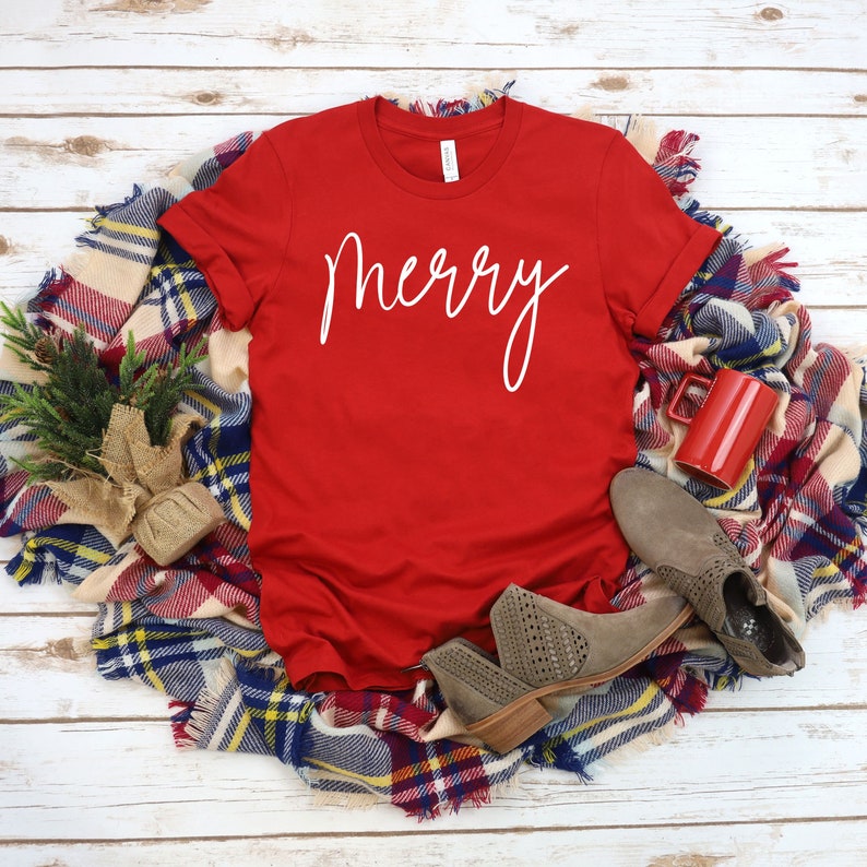 May include: Red t-shirt with the word "merry" written in white cursive lettering. The shirt is laying on a plaid blanket with a red mug and brown boots.