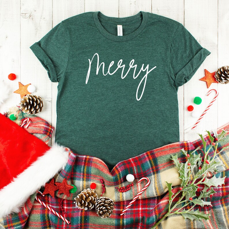 May include: A green t-shirt with the word "merry" written in white cursive on the front. The shirt is laying on a plaid blanket with red and white candy canes, pine cones, and holly.
