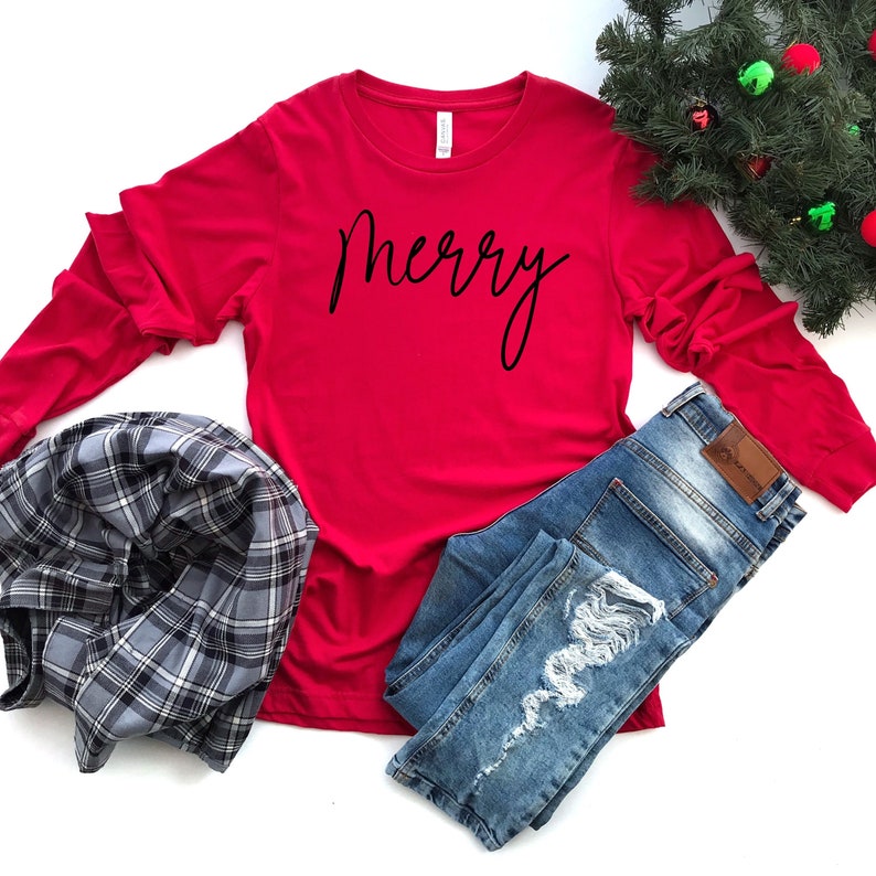 May include: Red long-sleeve shirt with the word "merry" written in black cursive script. The shirt is laid flat on a white background with a pair of blue jeans and a plaid shirt.