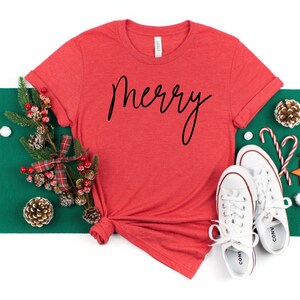 May include: Red t-shirt with the word "merry" written in black cursive script. The shirt is laid out on a white surface with a green and white background.