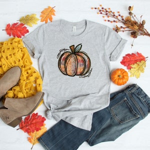 May include: A gray t-shirt with a colorful, hand-drawn pumpkin design. The pumpkin is surrounded by leaves and vines. The shirt is laid out on a white surface with fall-themed decorations.