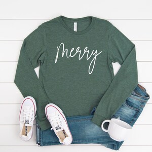 May include: A green long-sleeved shirt with the word "merry" written in white cursive on the front. The shirt is paired with blue jeans and white sneakers.