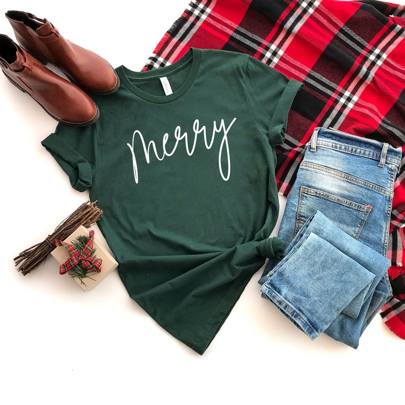 May include: A green t-shirt with the word "merry" written in white cursive script. The shirt is laid flat on a white background with a pair of brown boots, blue jeans, and a red and black plaid scarf.