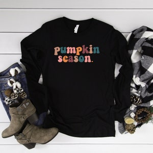 May include: Black long-sleeve shirt with the text "pumpkin season." in colorful, retro-style letters. The shirt is laid flat on a white wood surface with a pair of brown ankle boots and a black and white plaid scarf.