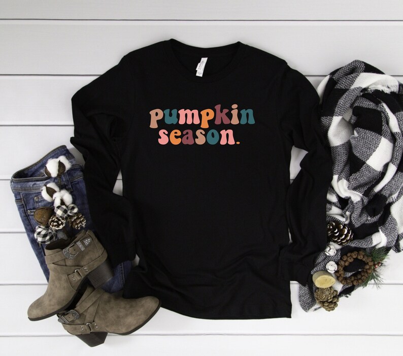 May include: Black long-sleeve shirt with the text "pumpkin season." in colorful, retro-style letters. The shirt is laid flat on a white wood surface with a pair of brown ankle boots and a black and white plaid scarf.