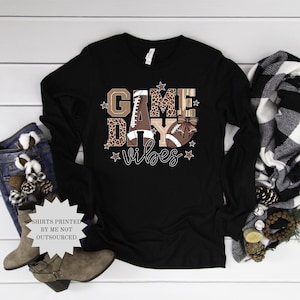 May include: Black long-sleeve shirt with a football-themed graphic. The design includes the words "Game Day Vibes" with a football and stars. The letters have a leopard print pattern.