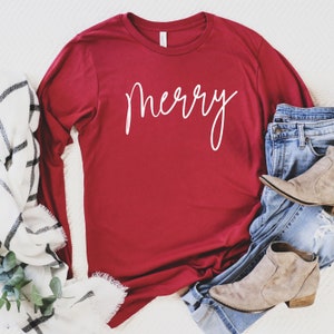 May include: A red long-sleeved t-shirt with the word "merry" written in white cursive script. The shirt is laid flat on a white surface with a white and black blanket and blue jeans with brown boots.
