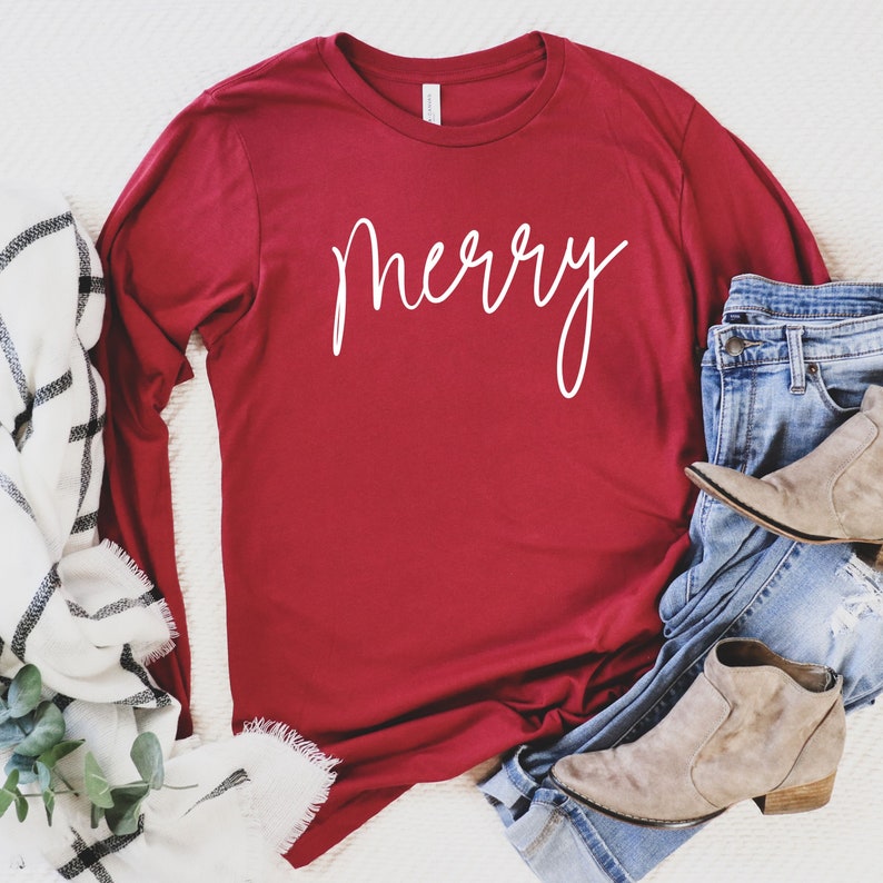 May include: A red long-sleeved t-shirt with the word "merry" written in white cursive script. The shirt is laid flat on a white surface with a white and black blanket and blue jeans with brown boots.