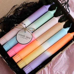 Könnte beinhalten: Ein Set mit sechs Ombre-Kerzen in einer Kartonbox. Die Kerzen sind in den Farben Rosa, Lila, Blau, Gelb, Orange und Grün. Die Box ist mit schwarzem Schredderpapier ausgekleidet. Die Kerzen sind mit "rumah saya studios" beschriftet.