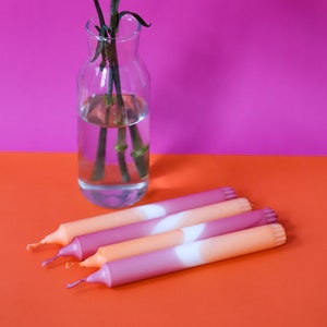 May include: Four ombre taper candles in shades of pink and orange with a white stripe in the middle. The candles are arranged on a bright orange surface.