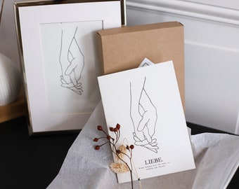 May include: A framed print of a line drawing of two hands holding each other. The print is on white paper and is framed in a silver frame. The print is sitting on a table next to a brown box with a white card that says "LIEBE.  Wenn Liebe das erste mal auf  anderer Person trifft."