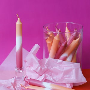 May include: A group of ombre candles in a glass jar, with one candle standing upright on a table. The candles are pink, white, and orange.