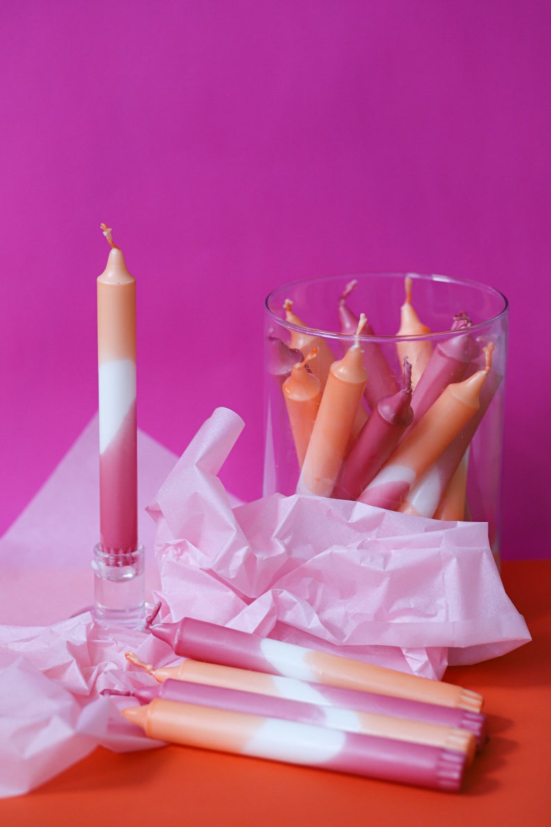 May include: A group of ombre candles in a glass jar, with one candle standing upright on a table. The candles are pink, white, and orange.