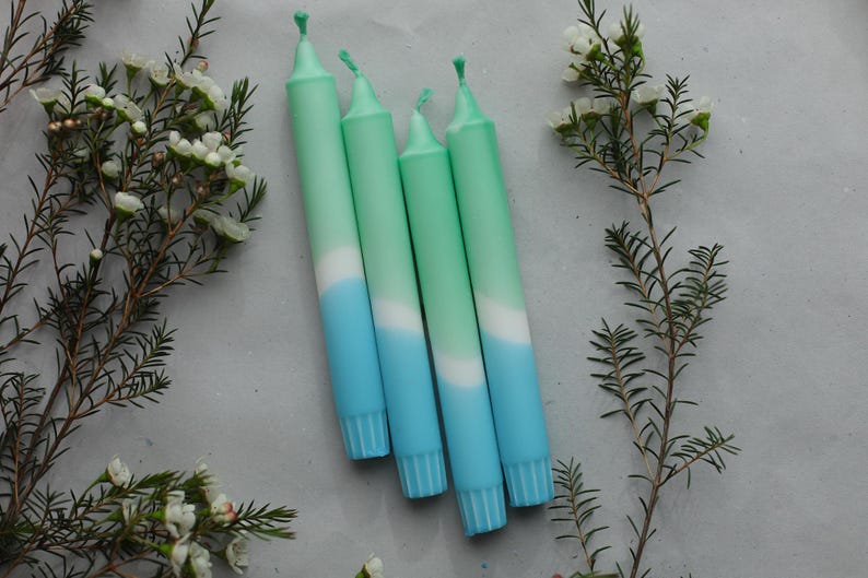 May include: Four ombre candles with a green to white to blue colour gradient. The candles are arranged in a row, with the green ends on the left and the blue ends on the right.