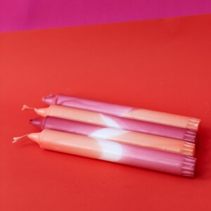 May include: Four pink and orange ombre candles with white accents. The candles are stacked on top of each other on a red surface.