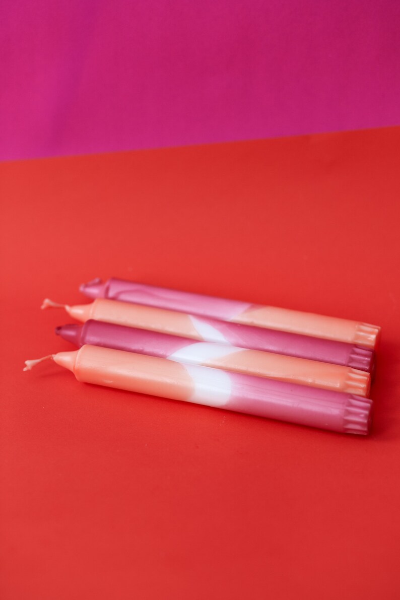 May include: Four pink and orange ombre candles with white accents. The candles are stacked on top of each other on a red surface.