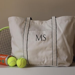 Könnte beinhalten: Eine cremefarbene Tragetasche mit schwarzen Zierleisten und den Initialen "MS". Ein Tennisschläger und eine Tube Tennisbälle liegen neben der Tasche. Die Tennisbälle sind gelb.