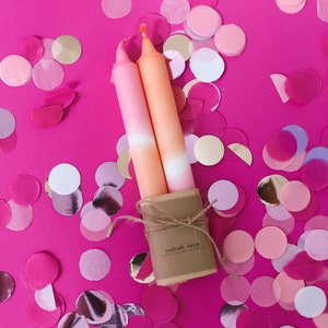 May include: Two ombre candles in pink and orange shades, tied together with twine and a brown paper label that reads "rumah saya". The candles are surrounded by pink, white, and gold confetti.