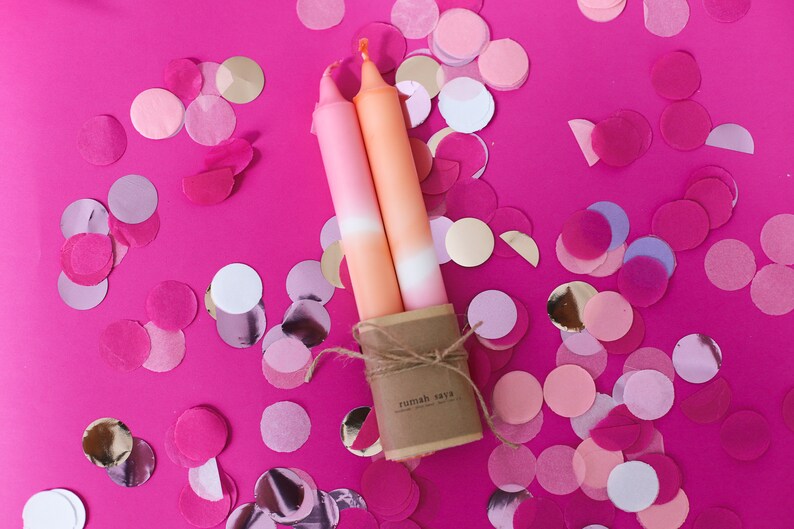 May include: Two ombre candles in pink and orange shades, tied together with twine and a brown paper label that reads "rumah saya". The candles are surrounded by pink, white, and gold confetti.