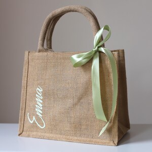 Peut inclure: Sac fourre-tout rectangulaire en toile de jute beige avec une poignée et un ruban vert clair noué en nœud. Le nom "Emma" est écrit en cursive sur le devant. Le sac est en matériau tissé.
