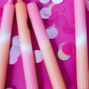May include: Four ombre pink and orange candles on a pink background with pink and white confetti.