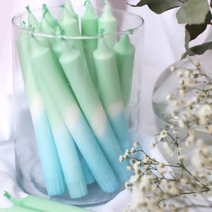 May include: A glass jar filled with ombre green and blue taper candles. The candles are arranged in a vertical stack and are all the same size. The jar is sitting on a white surface.