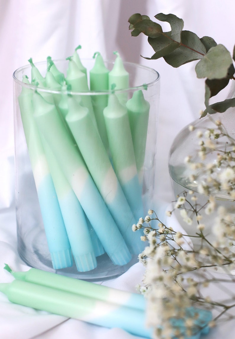 May include: A glass jar filled with ombre green and blue taper candles. The candles are arranged in a vertical stack and are all the same size. The jar is sitting on a white surface.