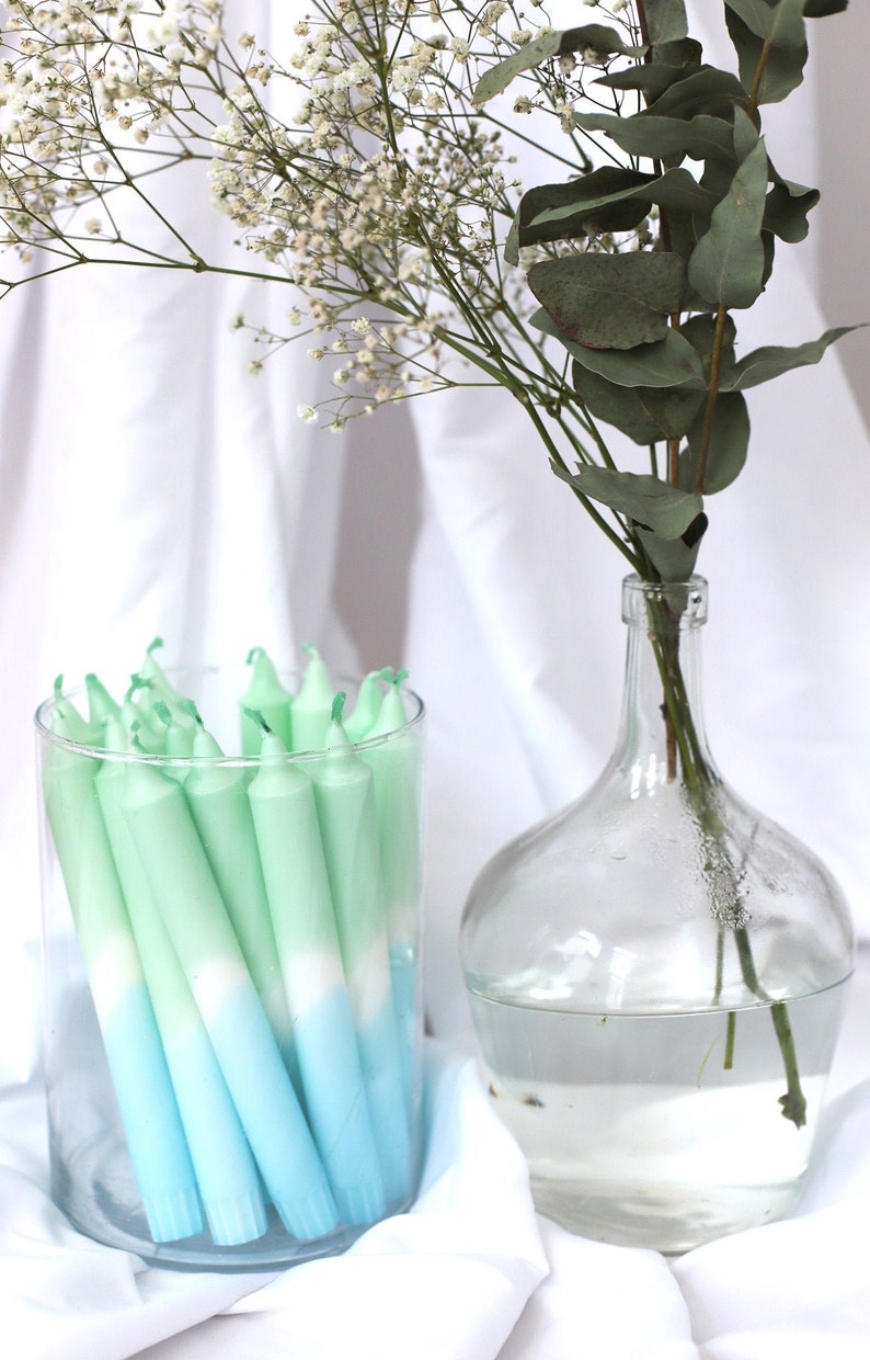 May include: A glass jar filled with ombre green and blue taper candles. The candles are arranged in a vertical stack. The jar is sitting on a white surface. A clear glass vase with a bouquet of white flowers and green leaves is in the background.