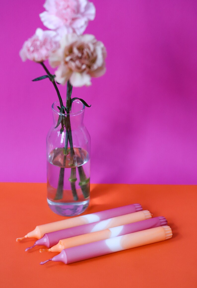 May include: Four ombre taper candles in shades of pink and orange with a white stripe in the middle. The candles are arranged on a bright orange surface.
