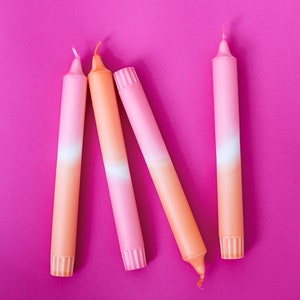 May include: Four ombre candles in shades of pink and orange. The candles are arranged in a row on a pink background.